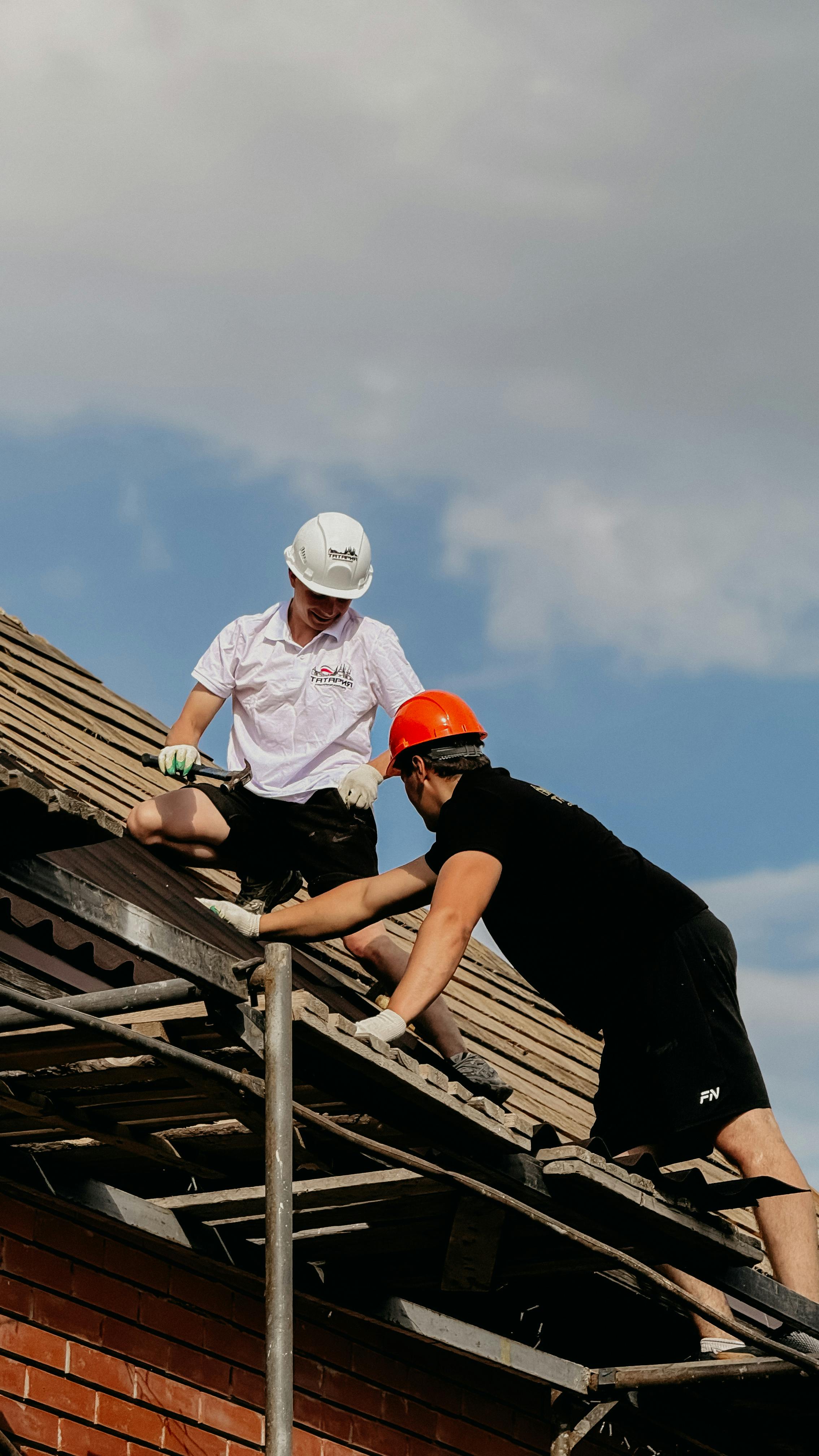Two workers in helmets are seen repairing a rooftop under a clear sky, emphasizing teamwork and maintenance.